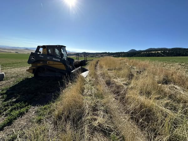 Compact track loader clearing and grading a dry field in Helena Montana under bright sun