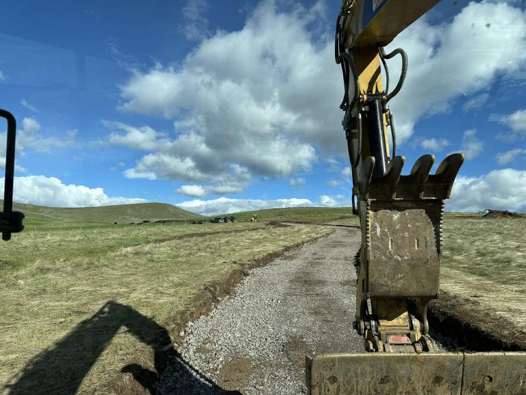 Excavator bucket shaping a gravel road through open fields under a blue sky in Montana