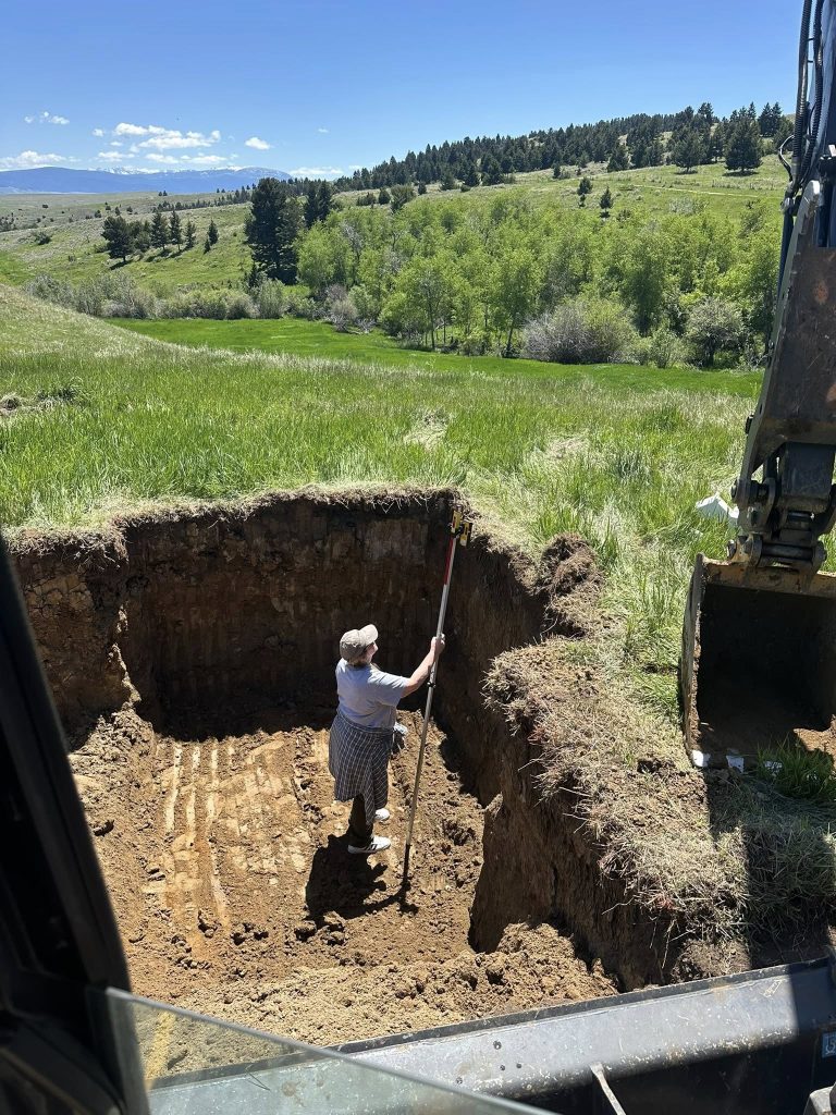 Excavation worker measuring a square foundation pit in a grassy Montana hillside