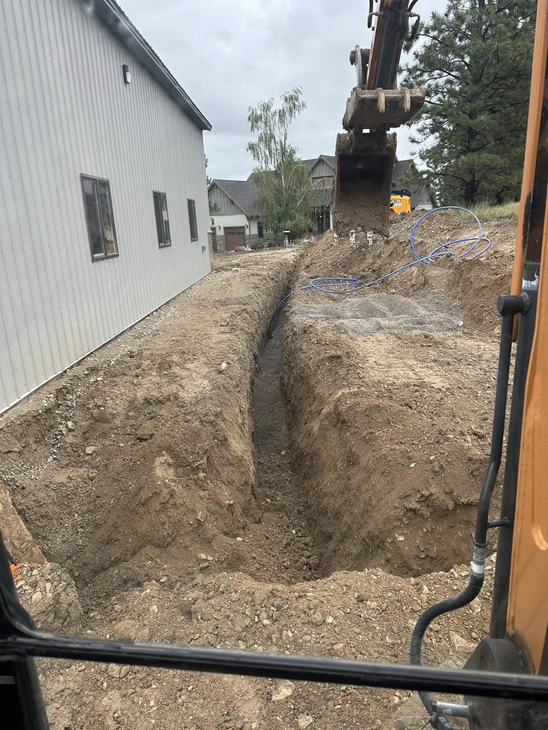 Excavator digging a precise trench along the side of a metal commercial building for utility line installation in Helena, Montana