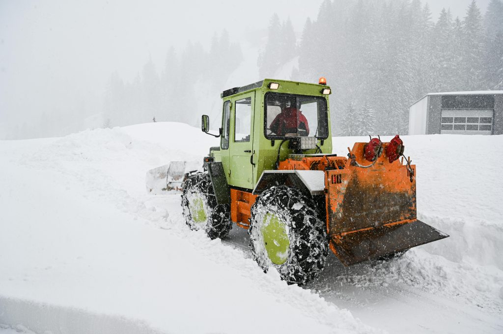 Green and orange snow plow truck clearing deep snow on a rural Montana road in heavy snowfall