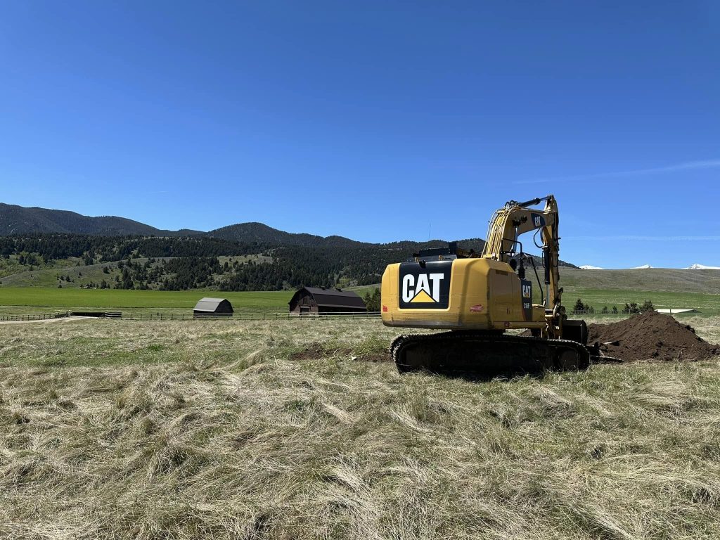 Yellow CAT excavator digging into a large open field during landscape and irrigation setup in Montana